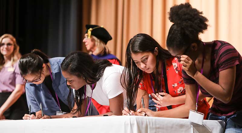 Four students lean on table to sign their names on the honor code.