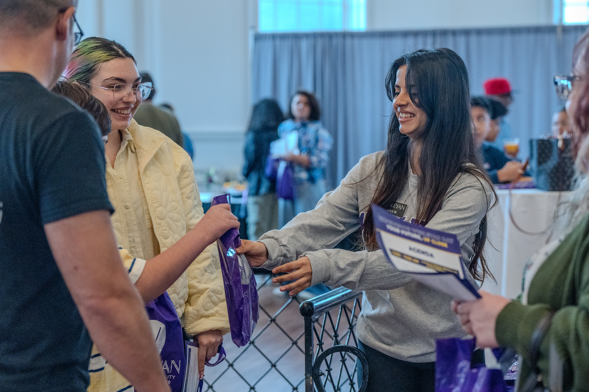 Ambassadors and Students Two young women smiling and interacting at an indoor event, as one hands a purple bag to the other. They are surrounded by other attendees in a bright, casual setting with tables and informational materials visible in the background.