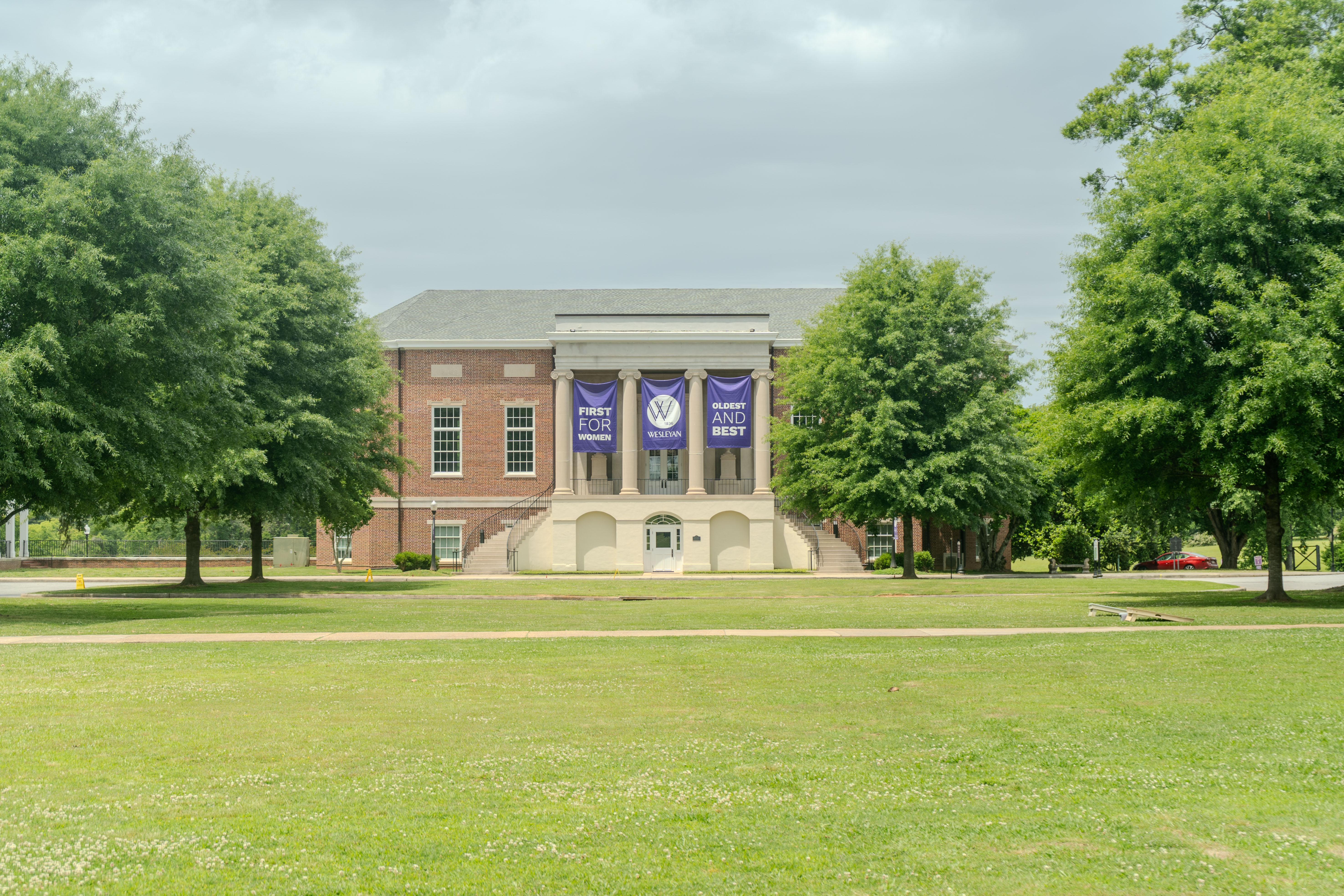 Candler from quad Front view of a stately brick campus building with white columns, centered across a green lawn and framed by large trees. Purple banners hang between the columns, including one with the Wesleyan logo and others reading “First for Women” and “Oldest and Be
