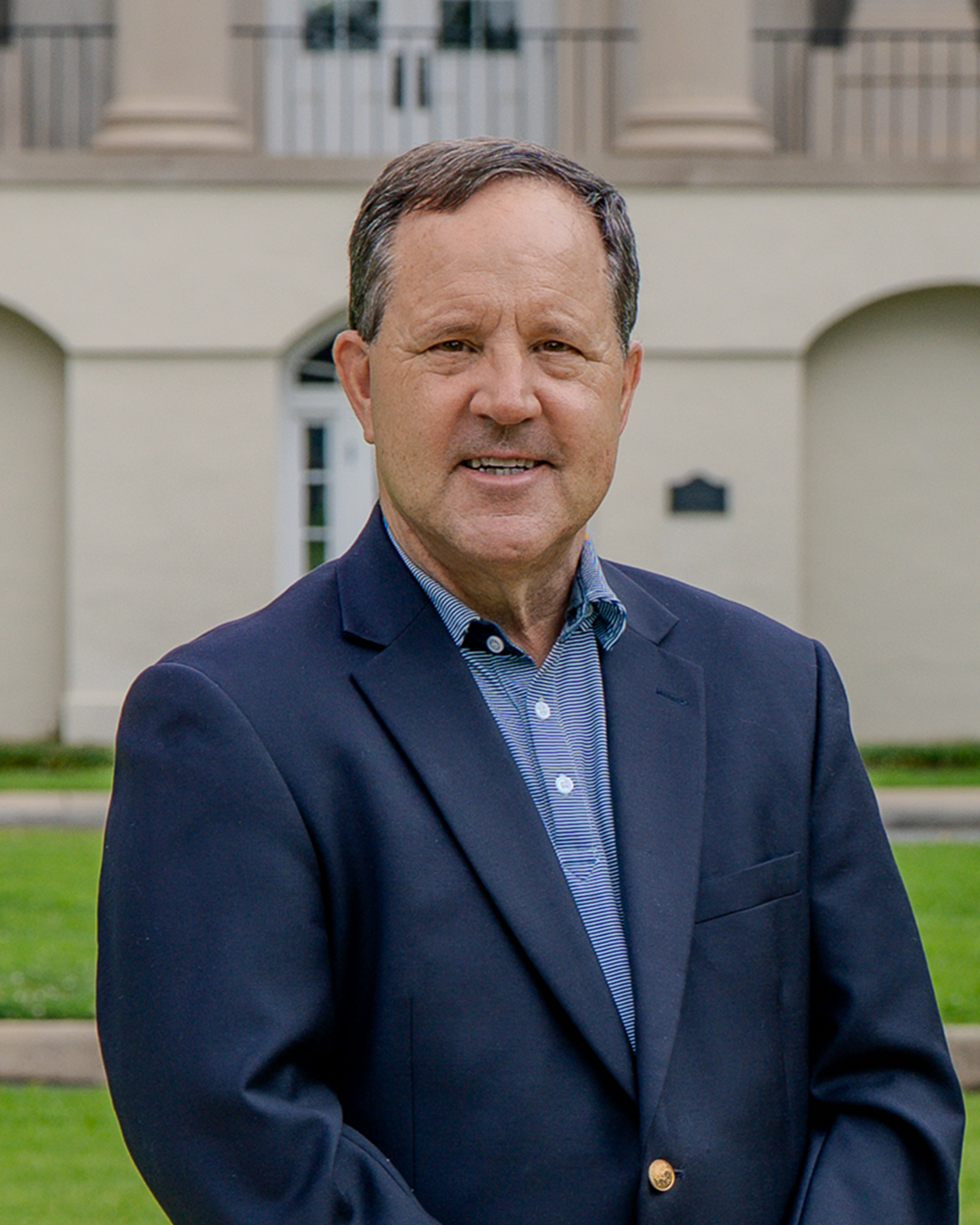 Middle-aged man wearing a navy blazer and striped button-down shirt, standing outdoors in front of a light-colored building with columns and balconies. He is smiling slightly and facing the camera, with a lawn visible in the foreground.